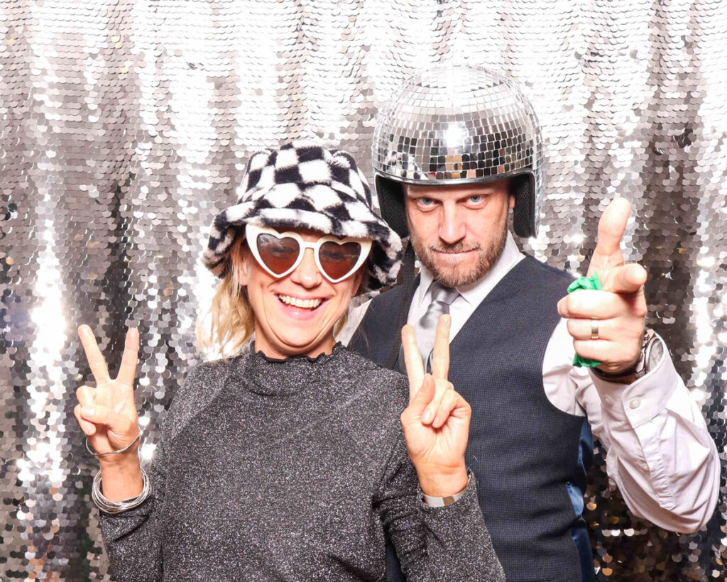 two people celebrating with festive props, hats, and a silver sequins photo booth backdrop during a corporate staff party entertainment