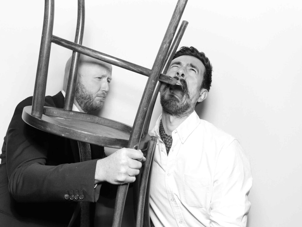 A close-up shot of two guests creatively using a chair in their photo booth prompt, showcasing their funny interaction against a white backdrop 