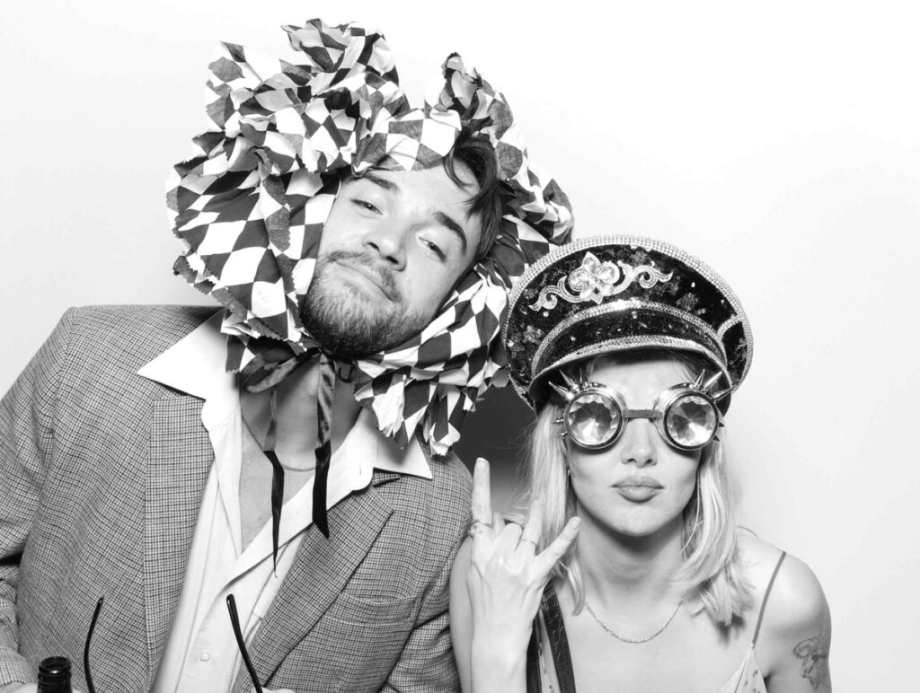 Two wedding guests posing in a photo booth, one wearing a playful ruffled headpiece and the other sporting a sequinned hat and steampunk-style goggles during a cripps barn wedding party entertainment

