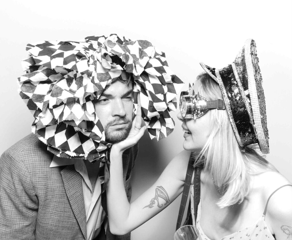 A humorous moment in the photo booth as one guest wearing a ruffled headpiece receives a playful cheek touch from another guest in a sequinned hat.