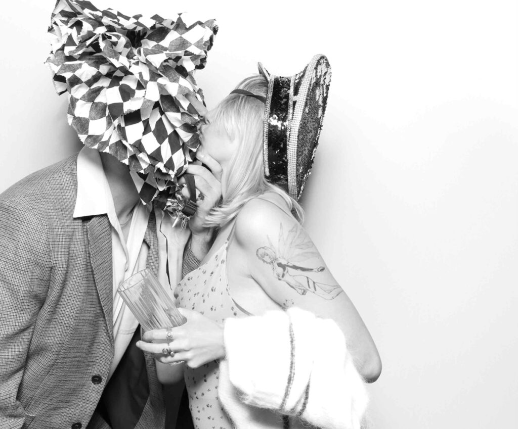 A fun and romantic photo booth moment with one guest in a ruffled headpiece sharing a kiss with another guest in a sequinned hat.