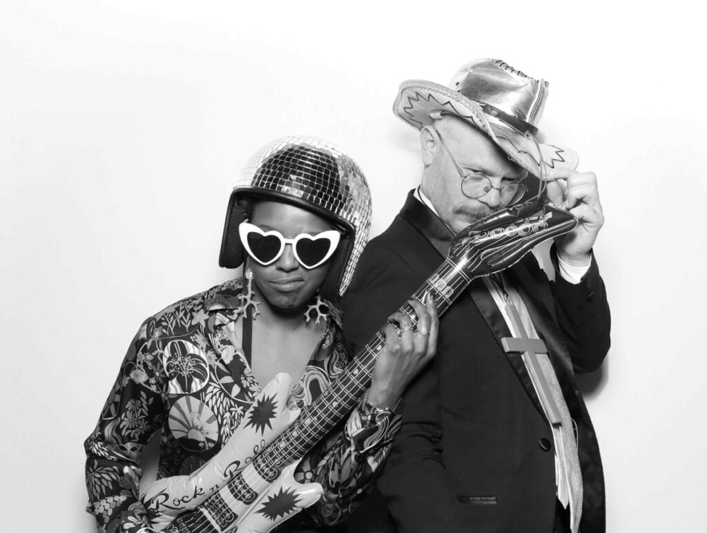 A joyful moment captured in black and white, with a guest in a disco helmet and another in a cowboy hat sharing a laugh in the photo booth during a cripps barn wedding entertainment