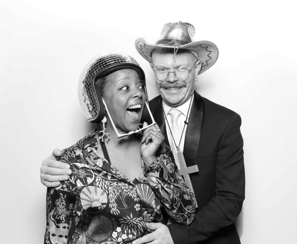 Two wedding guests in a photo booth with a musical theme, one wearing heart-shaped sunglasses and a disco helmet, the other tipping a cowboy hat.