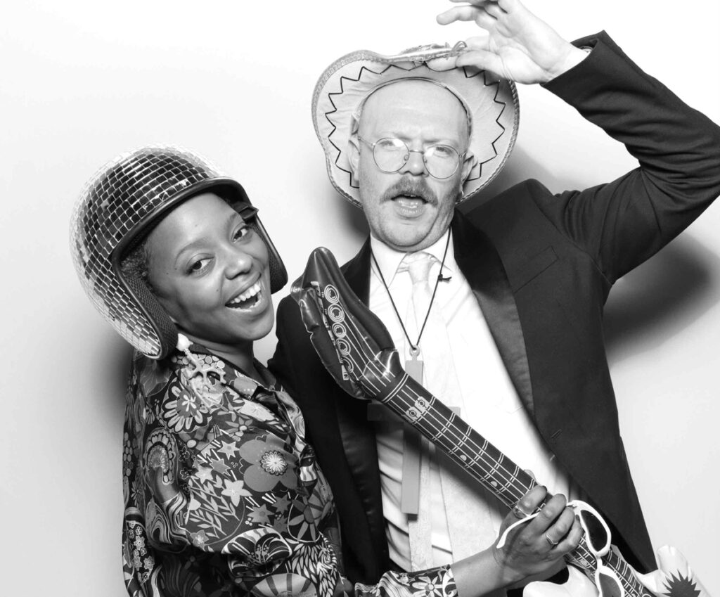 Wedding guests having fun with musical props in the photo booth, including an inflatable guitar and disco-themed accessories.