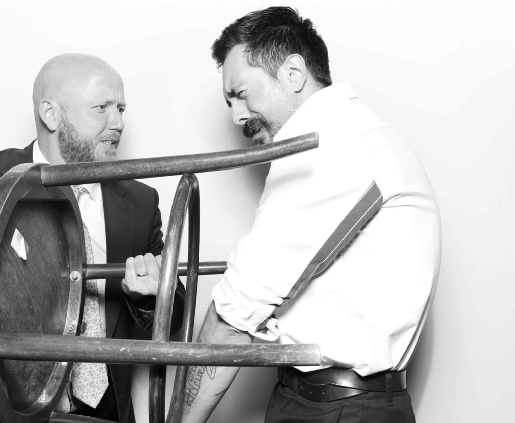 Two wedding guests in a photo booth acting out a humorous scene with a wooden chair, creating a playful moment in black and white.