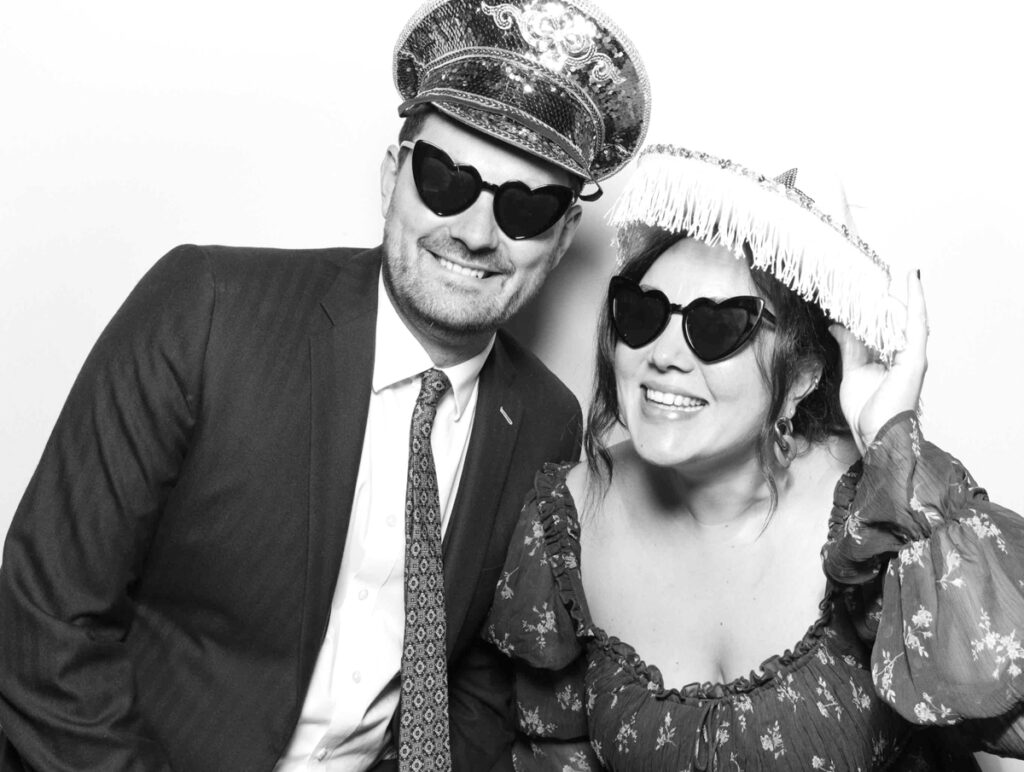 A cheerful photo booth picture at Cripps Barn featuring two guests smiling, both wearing heart-shaped sunglasses and festive hats.