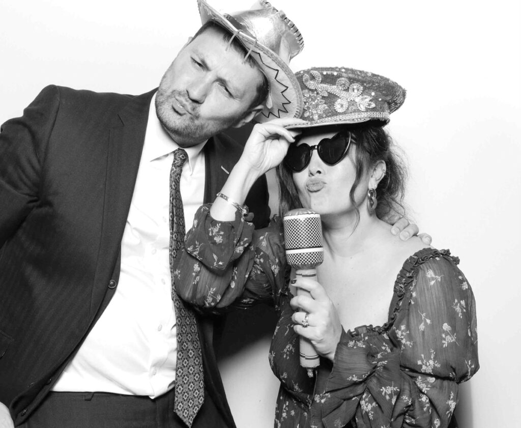 A playful pose in the Cripps Barn photo booth as guests wear sequinned hats and heart-shaped sunglasses, striking dramatic singing gestures with a prop microphone.