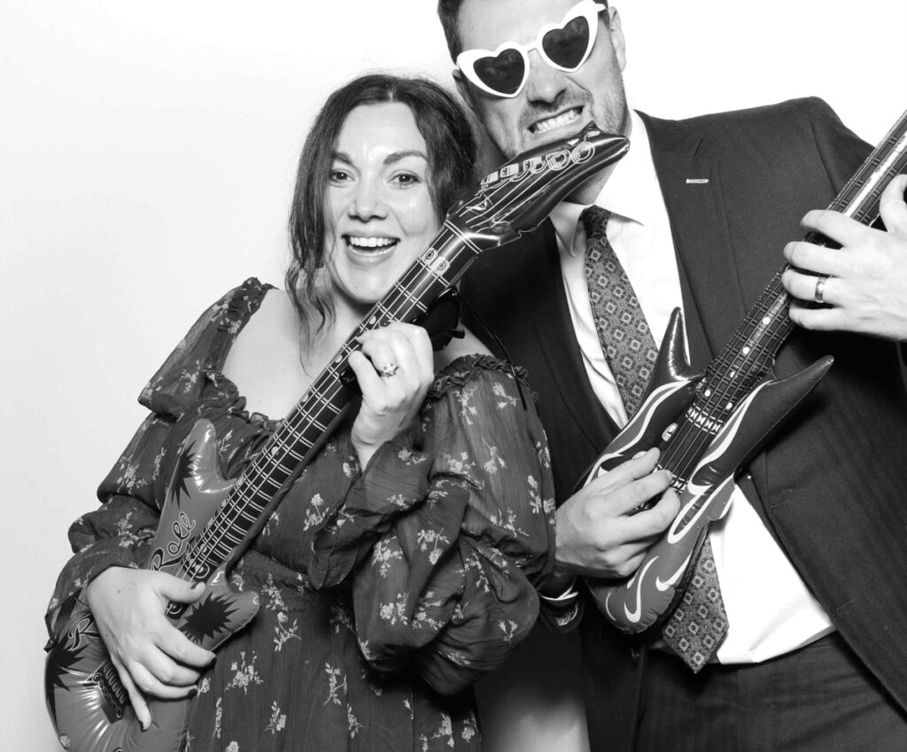 A fun photo booth moment at Cripps Barn with two guests enthusiastically playing inflatable guitars, one wearing heart-shaped sunglasses.