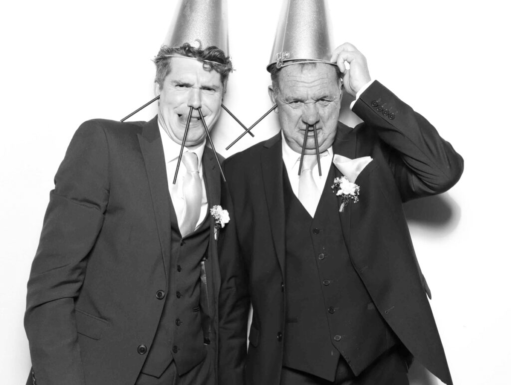 A hilarious moment in the Cripps Barn photo booth featuring two guests in suits with bucket hats and exaggerated facial expressions using straw props.