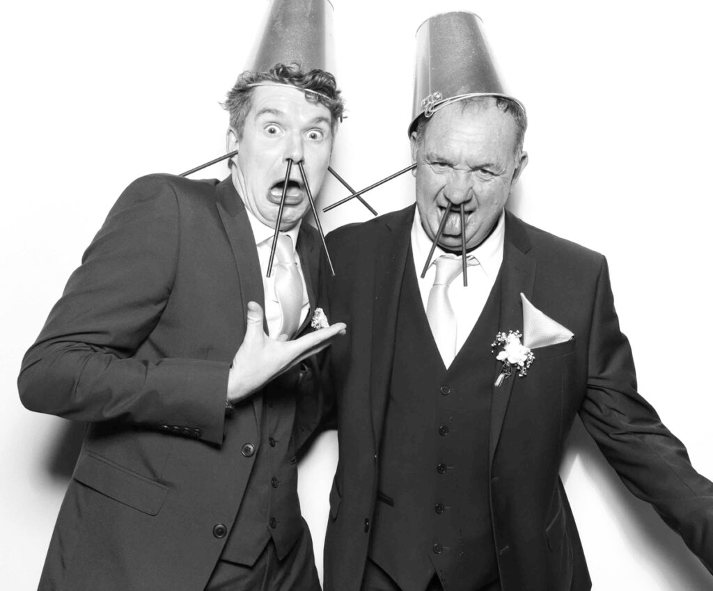 Two wedding guests at Cripps Barn posing in the photo booth with comical props, wearing buckets as hats and straws as walrus-like tusks.