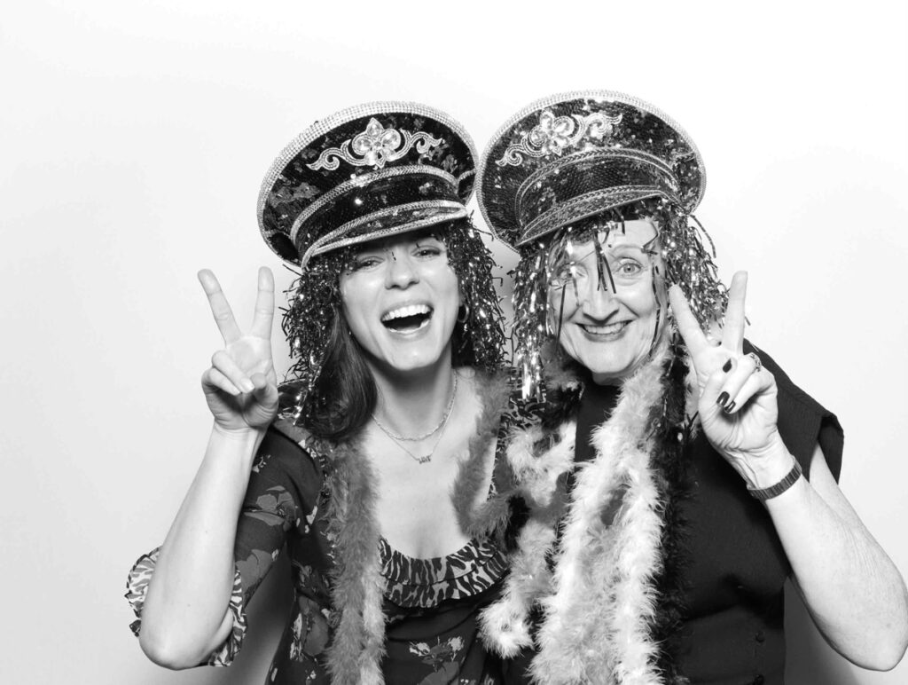 Guests at Cripps Barn sharing a warm and cheerful embrace in the photo booth, both wearing sparkling hats and surrounded by feather boas.