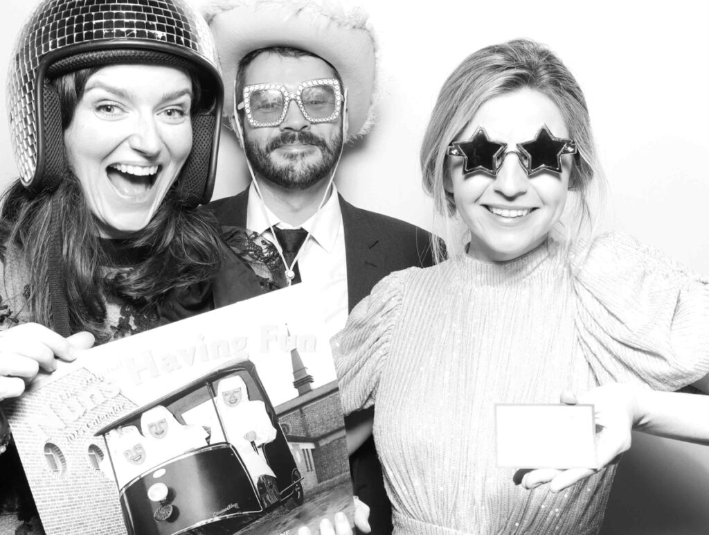 Three guests in the Cripps Barn photo booth, showcasing a mix of quirky props including a disco helmet, a cowboy hat, and star-shaped glasses, creating a joyful and lively atmosphere