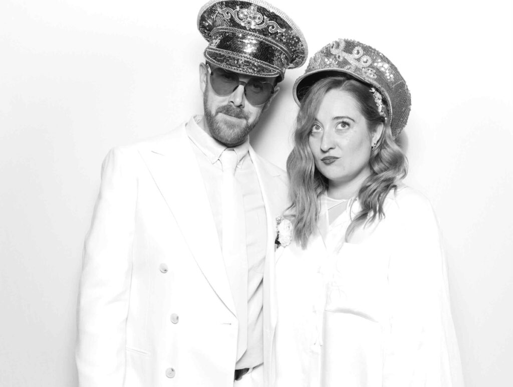 bride and groom in matching white outfits, wearing sequinned hats and posing confidently in the photo booth against a white backdrop for a kardashian style photo booth party entertainment