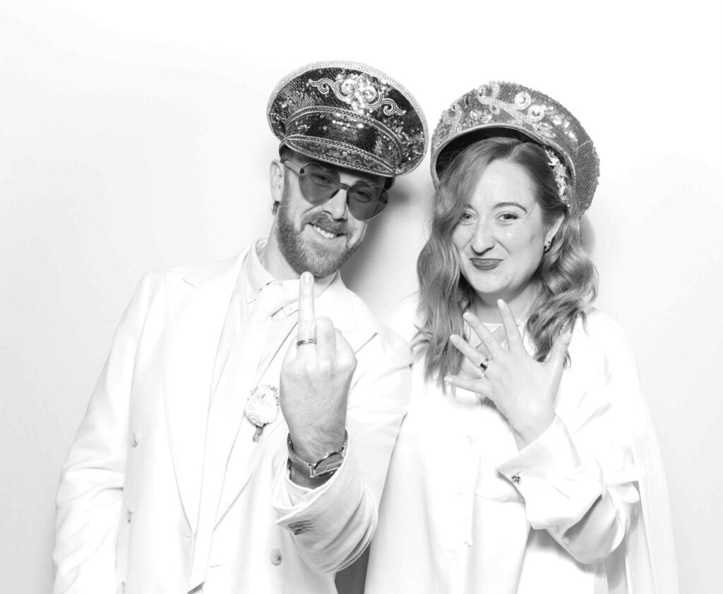 The couple striking a fun and energetic pose in the photo booth, both wearing matching sequinned hats and showcasing their wedding-day joy.