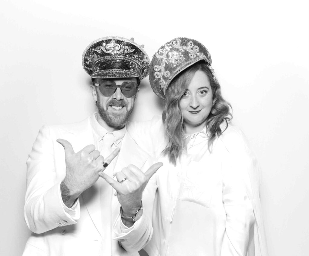 A playful photo booth moment featuring a couple in sequinned hats, one showing off their ring with a cheeky gesture and the other smiling warmly during a photo booth party entertainment