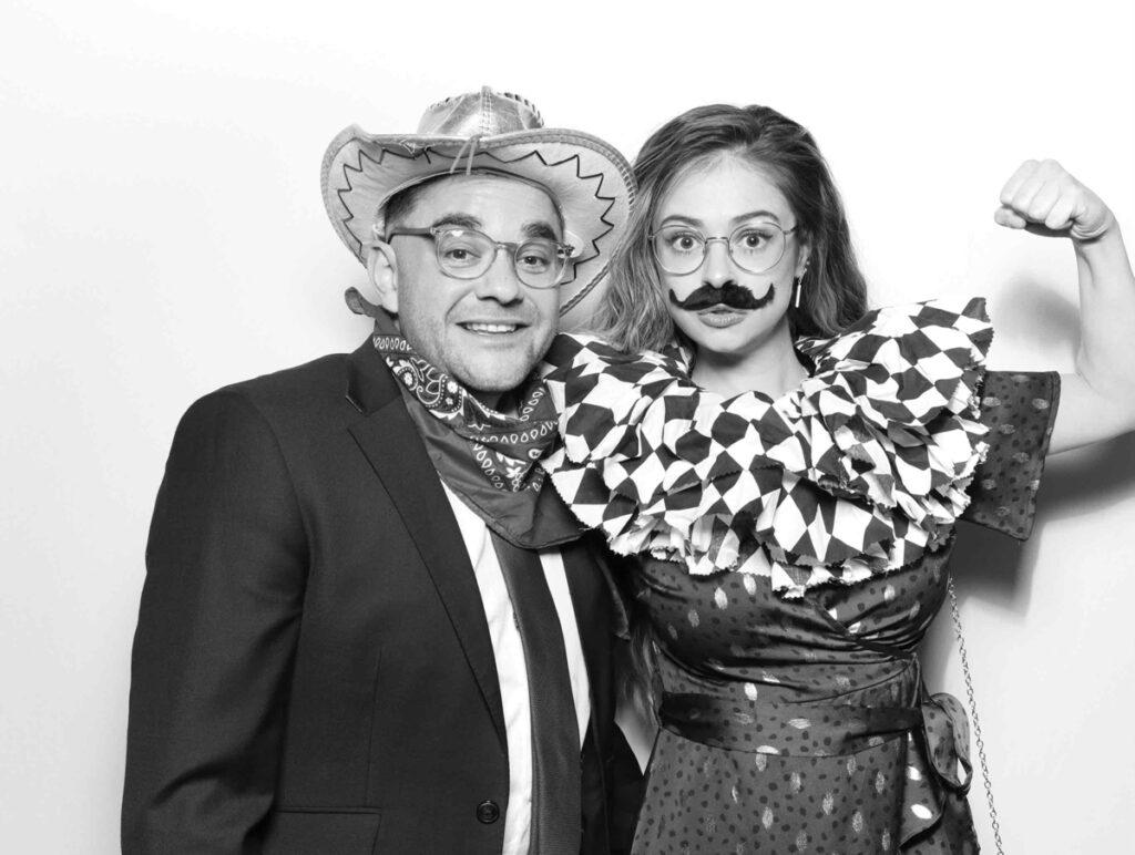 Two wedding guests posing in a photo booth, with one wearing a cowboy hat and the other playfully dressed with a moustache and ruffled costume.