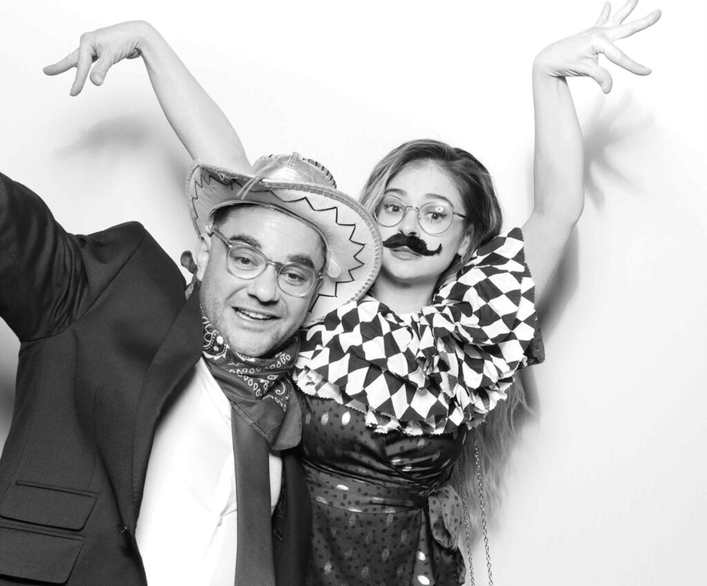 A playful photo booth moment featuring a guest in a cowboy hat smiling alongside a costumed guest with a fake moustache.