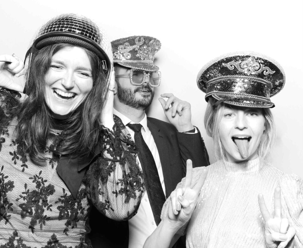 A vibrant photo booth moment at Cripps Barn featuring guests with heart-shaped sunglasses, cowboy hats, and feather boas, posing with big smiles and peace signs.