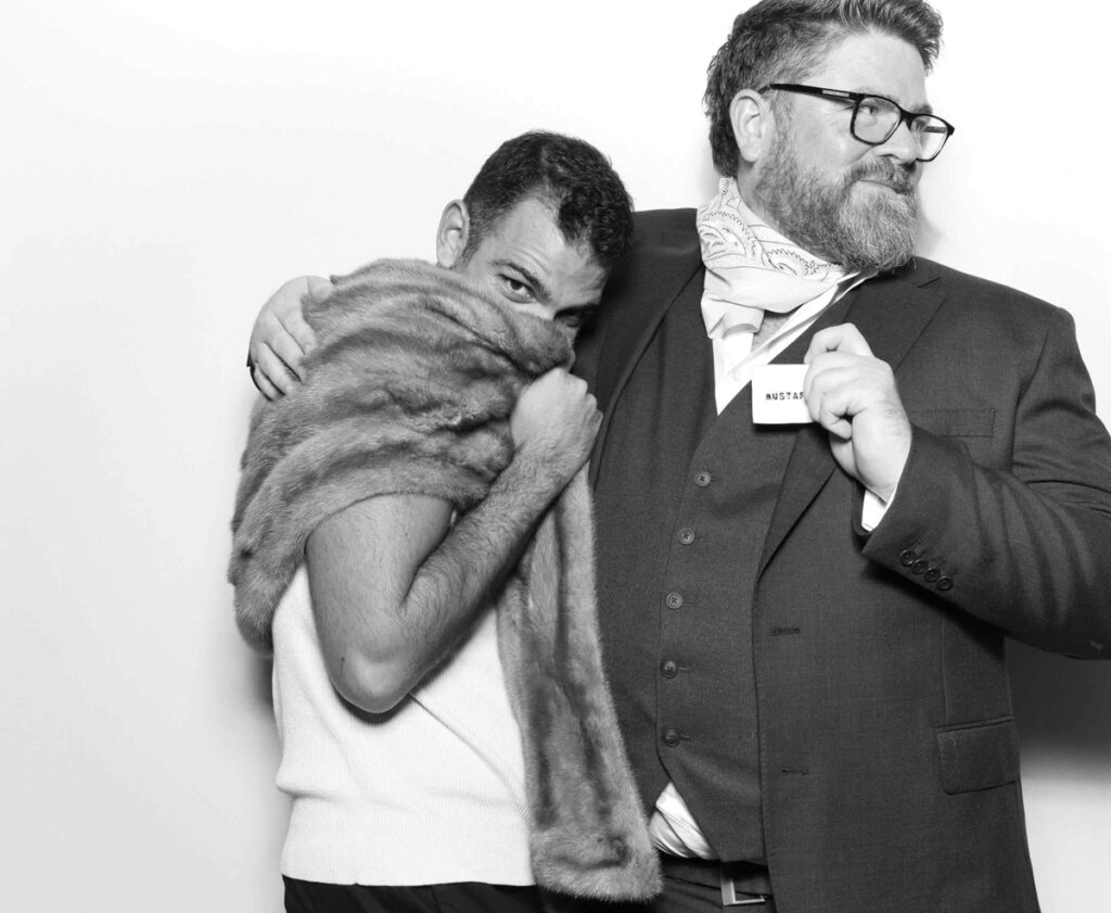 A playful shot of one guest draped in a fur stole and another posing with a name tag in the photo booth for the cripps barn wedding entertainment in the cotswolds 