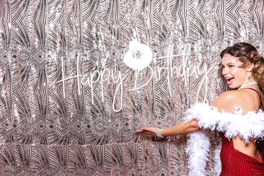 Smiling woman dressed in a glamorous red outfit and white feather boa posing against a gold-patterned photo booth backdrop with 'Happy Birthday' signage during a 1930s birthday party event entertainment