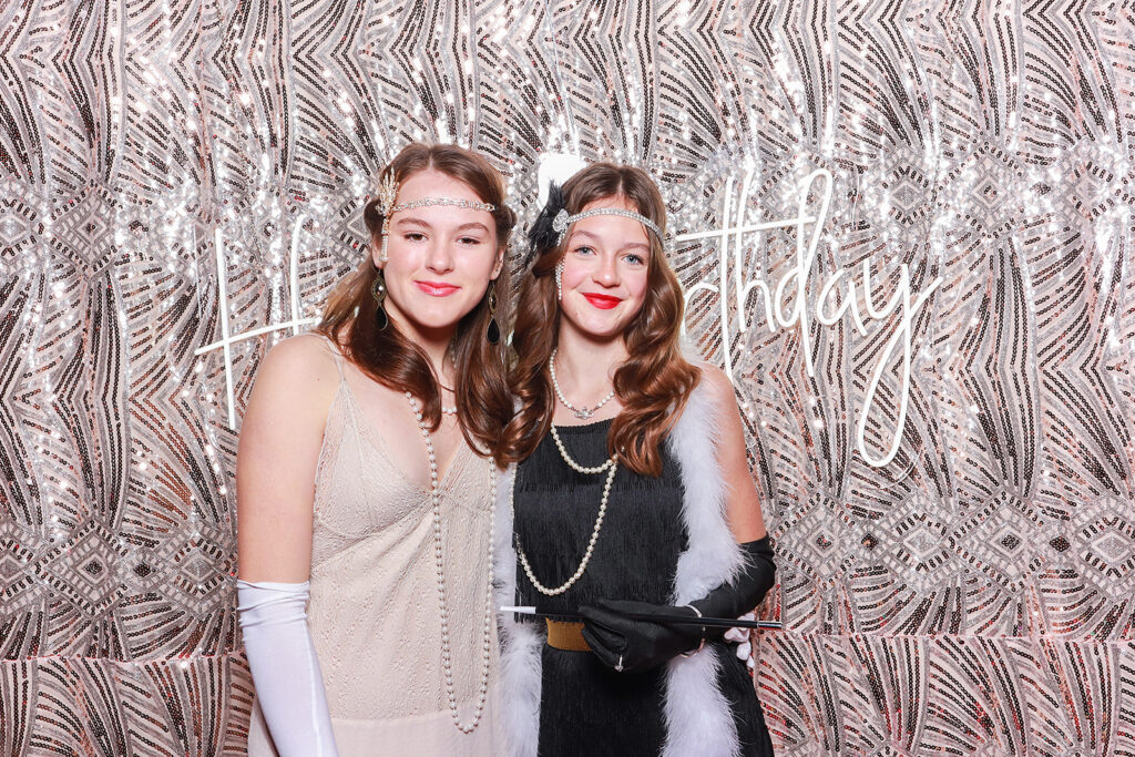 Two girls dressed in 1920s-inspired outfits with elegant headbands, pearls, and gloves, posing in a photo booth with a 'Happy Birthday' sign and a gold-patterned backdrop at a birthday party