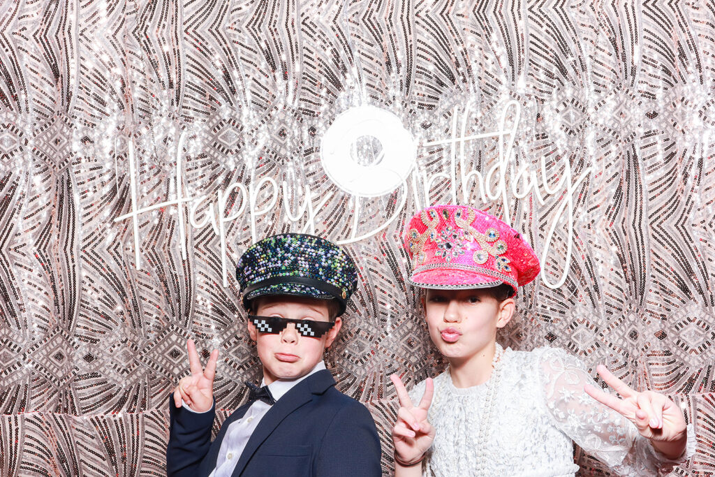 Two children dressed in stylish outfits posing with playful hats and novelty glasses in front of a 'Happy Birthday' sign and a gold-patterned photo booth backdrop at a birthday party in Oxfordshire