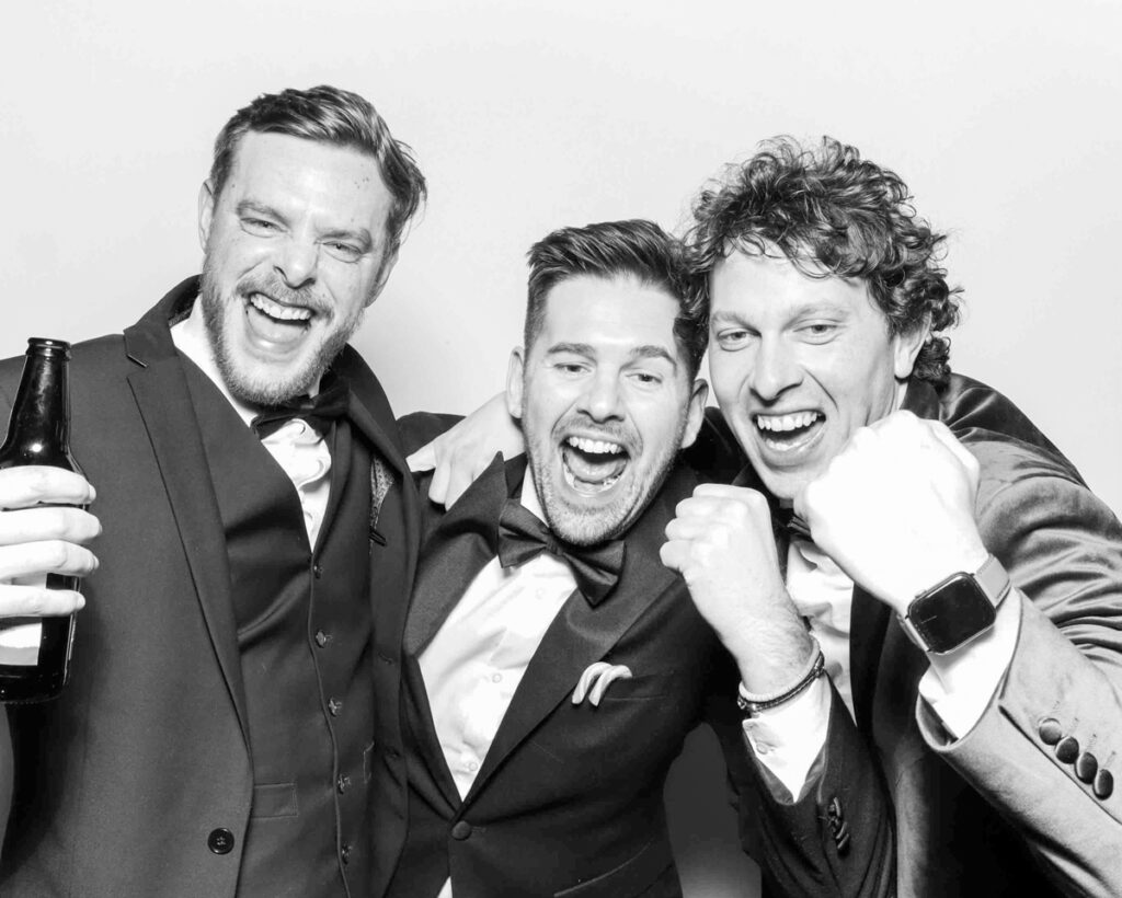 Three men dressed in black tie, cheering and laughing in a black and white photo booth session at a corporate event or wedding in Oxfordshire