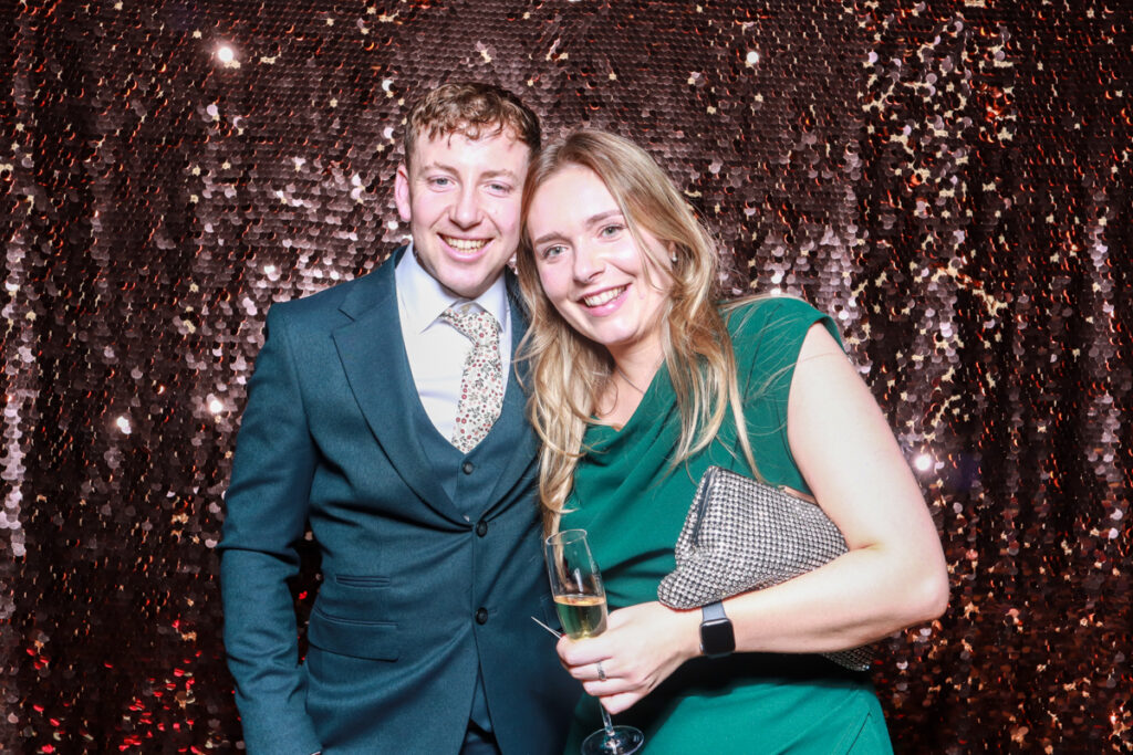 Smiling couple dressed in formal attire posing with a glass of champagne in front of a sparkling sequin backdrop at a christmas staff party entertainment