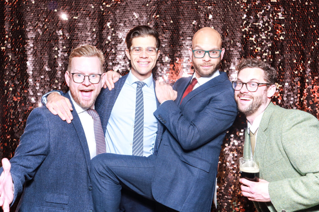 Four men in suits posing playfully in front of a shimmering sequin backdrop, captured during a corporate event or party in Oxfordshire