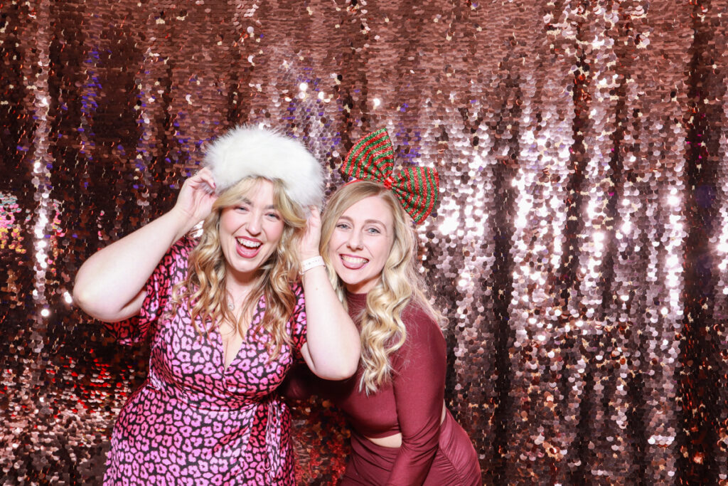 Two women posing with festive accessories, including a furry white hat and a red and green bow, against a sparkling sequin backdrop at a Christmas party or event 