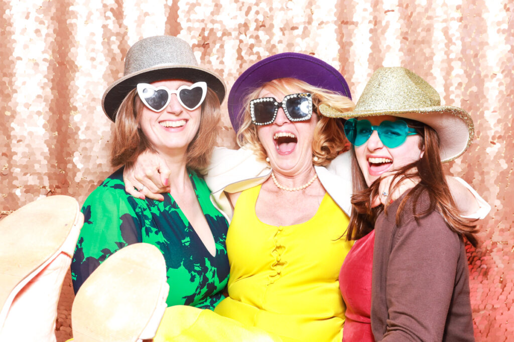 Three women laughing and enjoying themselves in a photo booth, wearing vibrant outfits and stylish hats with heart-shaped and square sunglasses, against a sparkling gold backdrop at a birthday party event