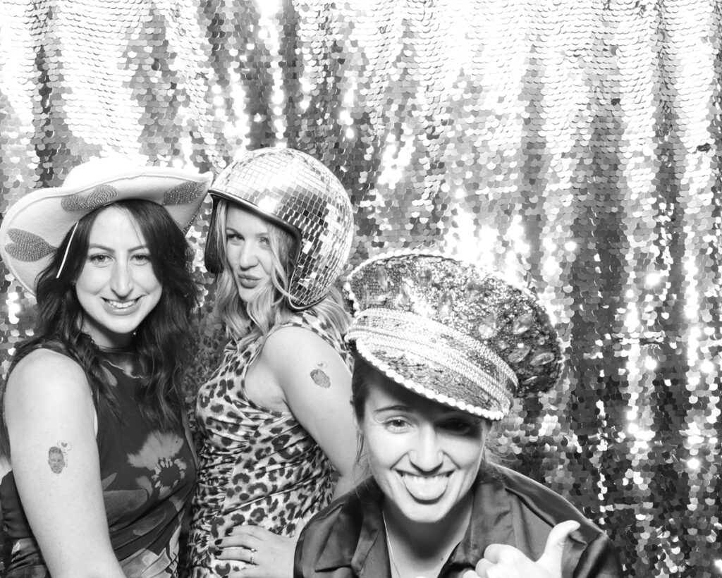 Three women posing in a black and white photo booth with fun accessories, including a cowboy hat, disco ball helmet, and a sparkly captain's hat, against a shimmering sequin backdrop at a corporate event 