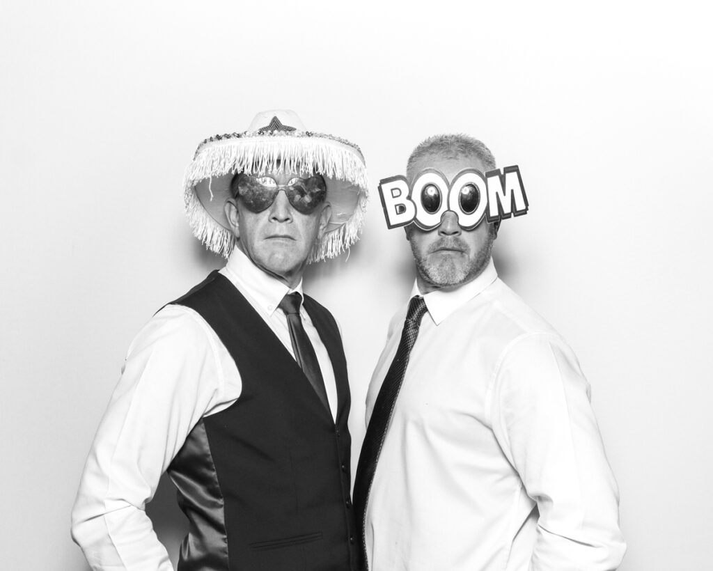 Two men in formal attire posing with playful props, including a fringe cowboy hat and novelty 'Boom' glasses, during a black and white photo booth session at an Oxfordshire event