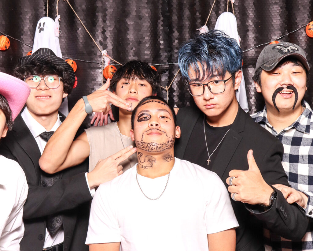 A group of young men posing with Halloween-themed decorations and props, including drawn-on face art and fun accessories, at a photo booth event i