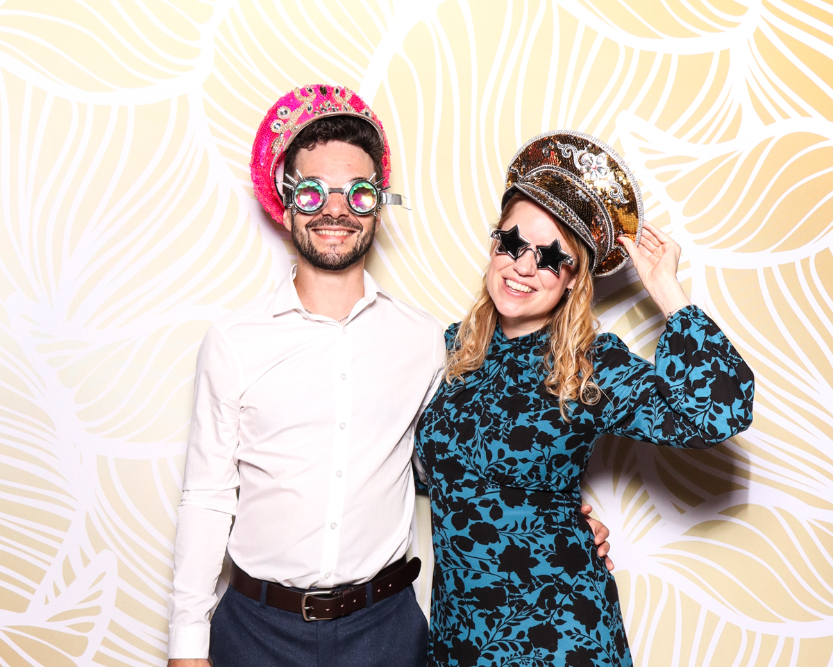 A young man and woman smiling together in front of a modern, artistic gold and white leaf-patterned backdrop. The woman is holding a novelty mask shaped like a dog's face wearing a top hat.