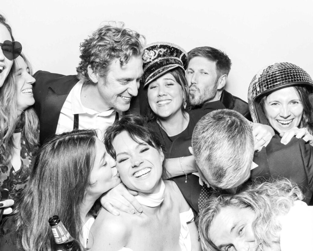 A lively group of guests laughing and embracing while posing with fun props, including a sequinned captain's hat and disco ball helmet, in a black and white photo booth session at a wedding or event in Oxfordshire.