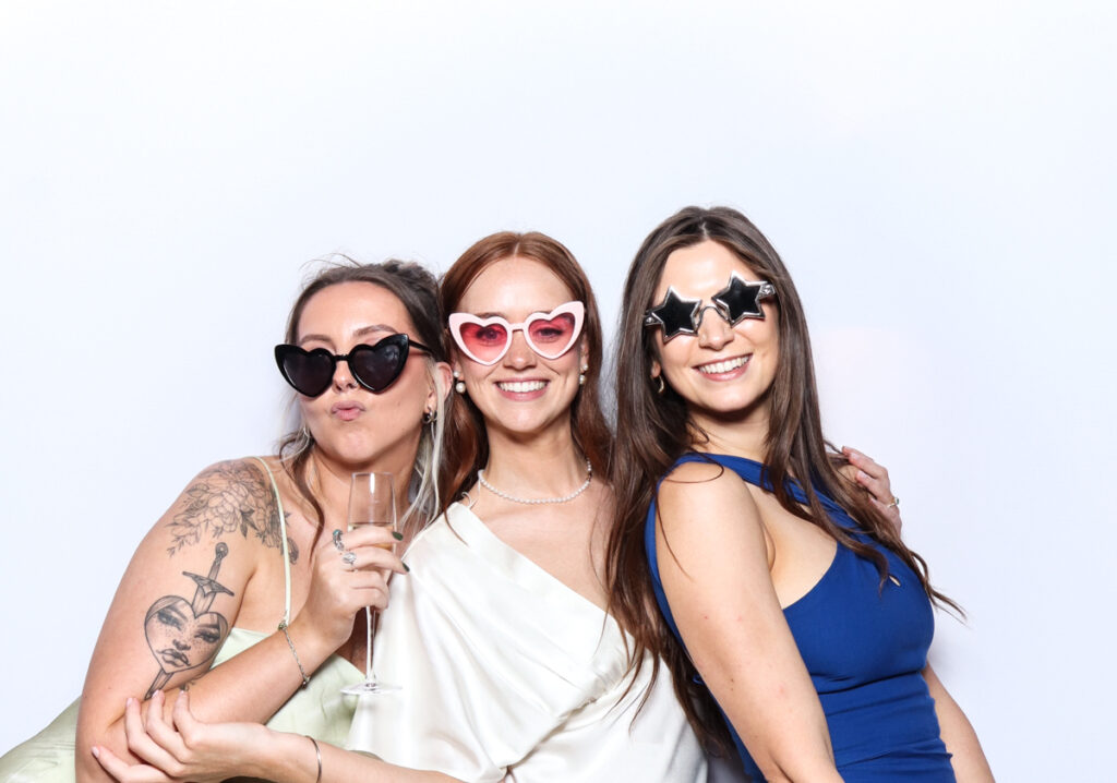 Three women posing in a photo booth wearing fun heart-shaped and star-shaped sunglasses, with one holding a champagne glass, against a white backdrop during a high end event near Cotswolds