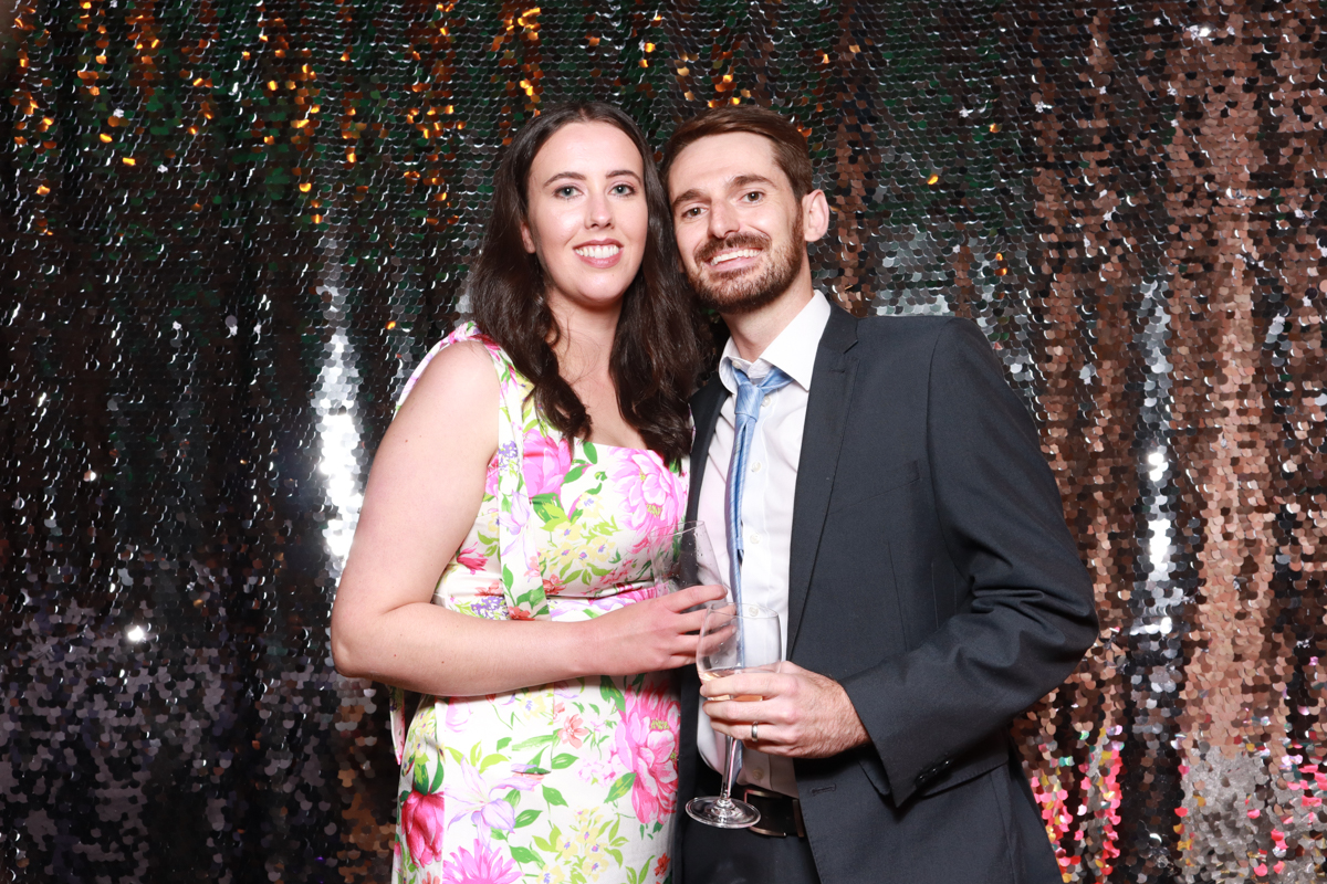 A couple dressed elegantly posing in front of a sparkling copper sequin backdrop, holding champagne glasses and smiling warmly at the camera.