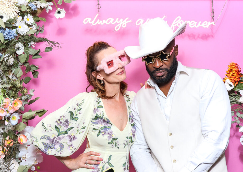 A stylish couple posing in a photo booth with a pink floral backdrop, wearing playful props including fluffy pink glasses and star-shaped sunglasses, with a neon 'always and forever' sign at a wedding or event in Oxfordshire