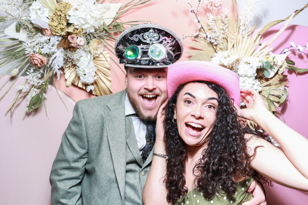 a fun image of a smiley couple posing against an arched backdrop with dried flower arrangement during the wedding reception photo booth hire in oxfordshire