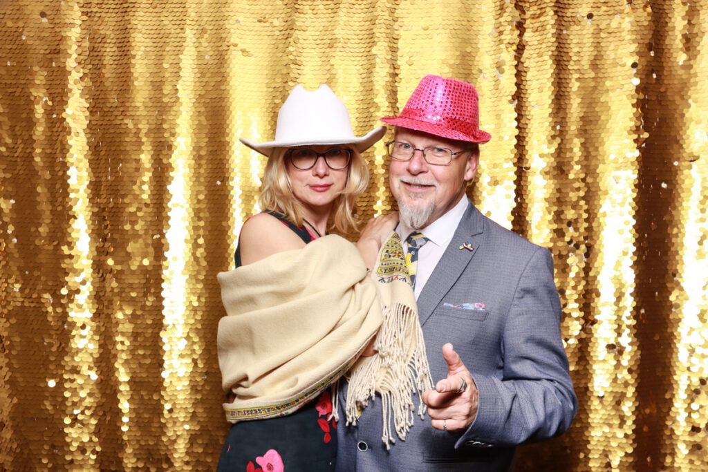 an older couple elegantly dressed,, wearing fun fats, posing against a golden sequins backdrop, during a corporate event near Cotswolds