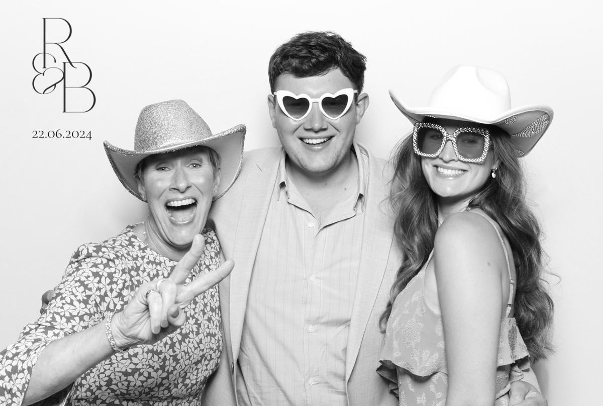 Three smiling guests posing in a black and white photo booth, wearing cowboy hats and heart-shaped sunglasses, with a monogram and date '22.06.2024' visible in the corner, at a weddin