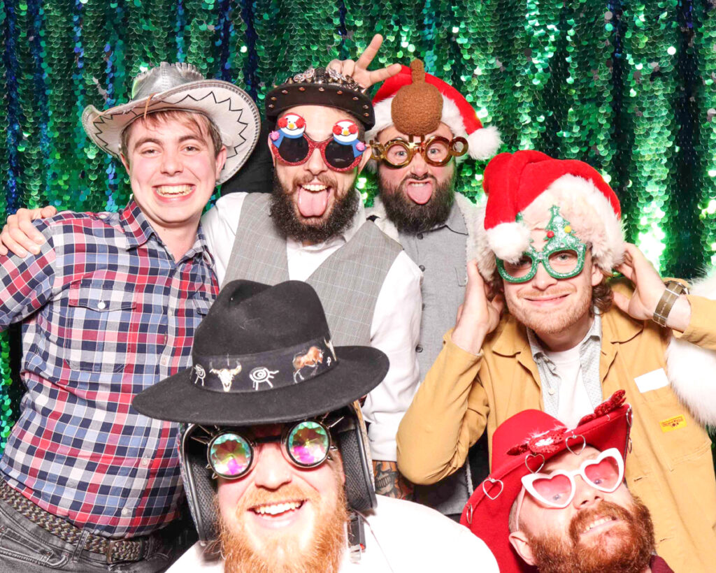 Group of guests in fun festive props, including Santa hats, novelty glasses, and cowboy hats, posing in a photo booth with a sparkly green backdrop at a Christmas party in Oxfordshire.