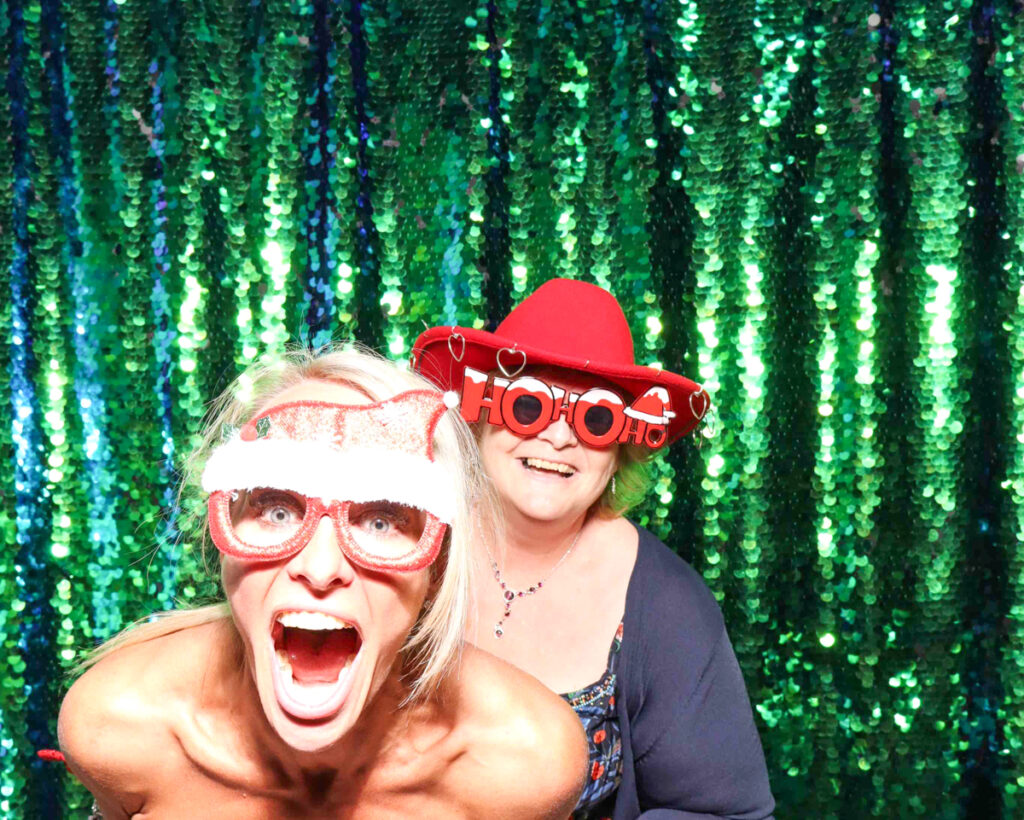 Two women posing in a festive photo booth with a green sequin backdrop, wearing fun Christmas-themed props, including a Santa hat, novelty glasses, and a red cowboy hat that says 'Ho Ho Ho', during a Christmas party in Oxfordshire.