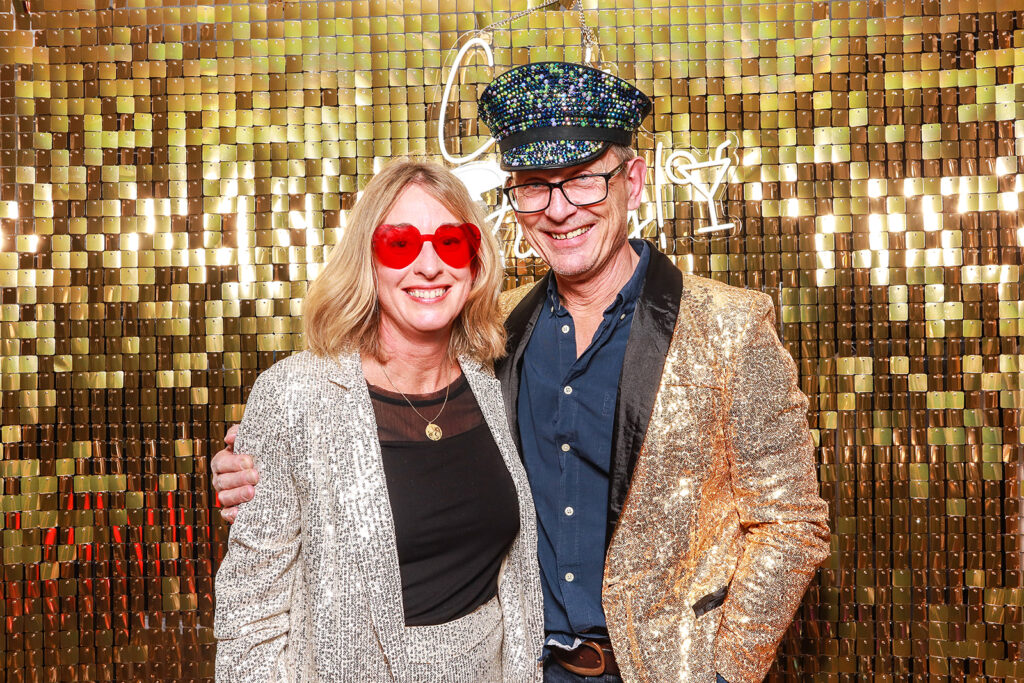 Smiling couple dressed in sparkling outfits, with red heart-shaped sunglasses and a sequinned captain’s hat, posing in front of a shimmering gold photo booth backdrop at a brand opening event in Oxfordshire