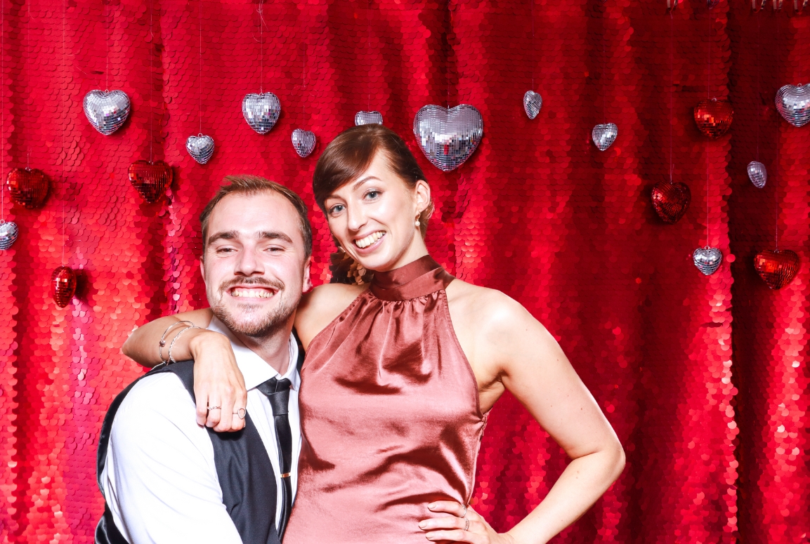 couple posing against a red sequins backdrop with disco ball heart - shaped props hanging against the backdrop during a valentines day photo booth event, the perfect entertainment for shopping malls and restaurants