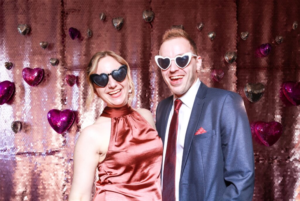 couple posing against a lilac sequins backdrop with disco ball heart - shaped props hanging against the lilac sequins backdrop during a valentines day photo booth event, the perfect entertainment for shopping malls and restaurants