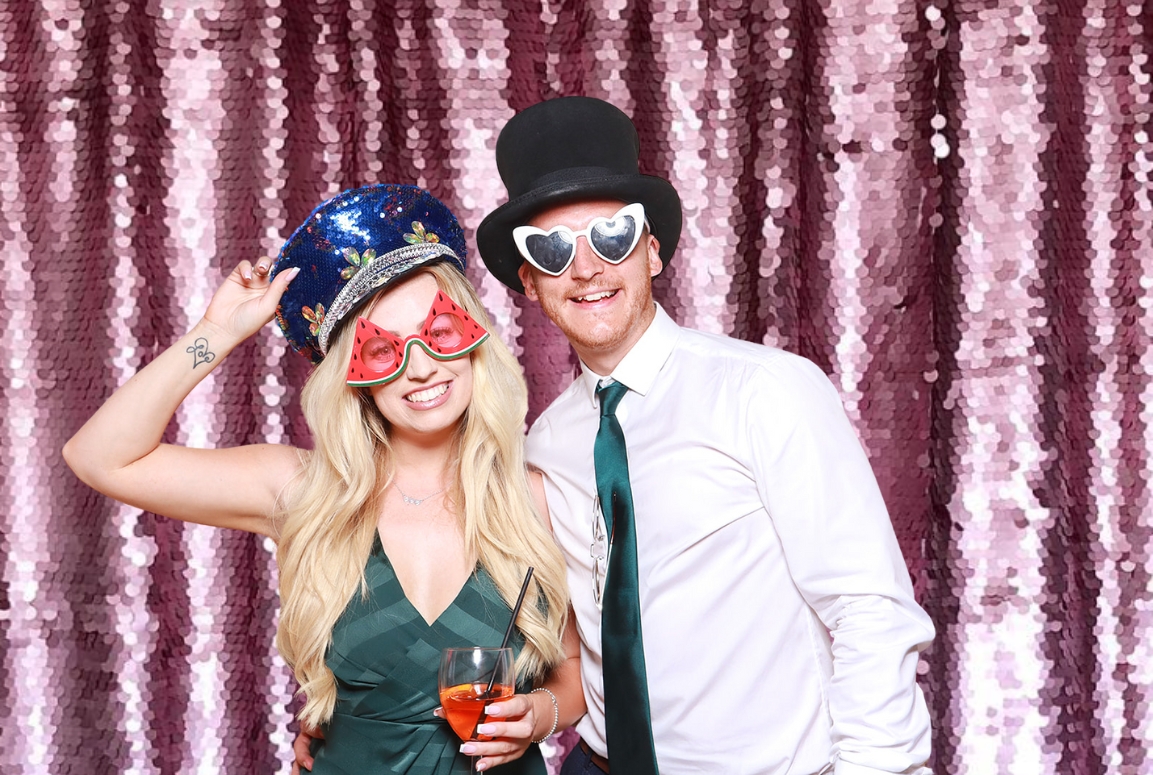 couple posing against a lilac sequins backdrop wearing fun props and heart shaped sunglasses, during a valentines day photo booth event, the perfect entertainment for shopping malls and restaurants 