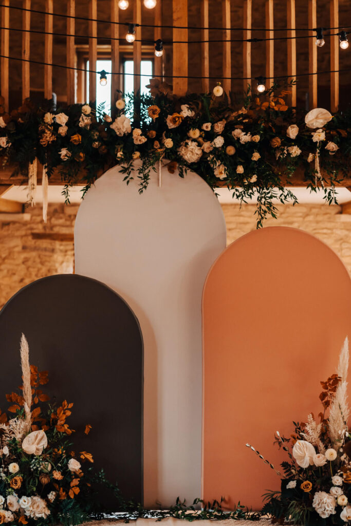 A stylish wedding ceremony backdrop featuring three modern arched panels in neutral tones, decorated with lush floral arrangements and warm string lights in a rustic barn venue.