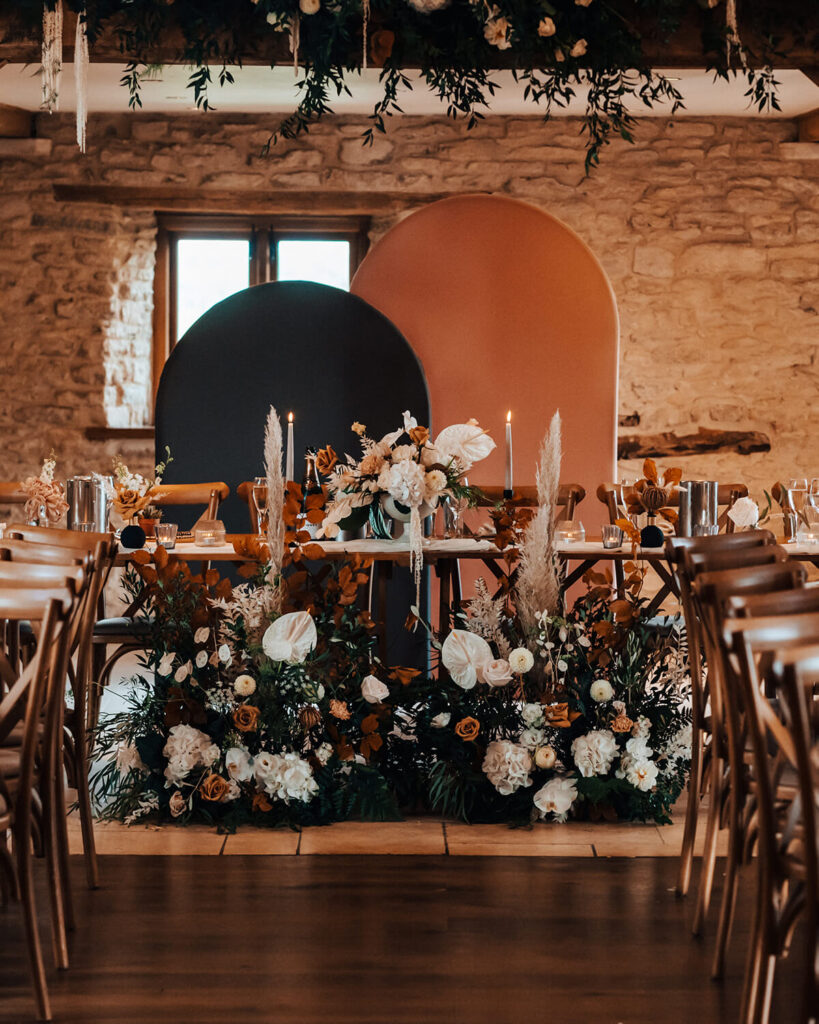 A beautifully styled wedding reception setup with floral-adorned arched backdrops in warm, earthy tones, complemented by candlelit tables and rustic wooden chairs in a cozy barn setting